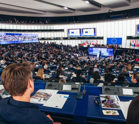 A large assembly hall filled with people attending a formal event. The room has a modern, semi-circular seating arrangement with individual desks equipped with microphones. A speaker stands at a podium addressing the audience, with large screens displaying the European Union (EU) logo and text. Several EU flags are visible in the background, along with a stage featuring an orchestra. Attendees are engaged in listening, taking notes, and using electronic devices.